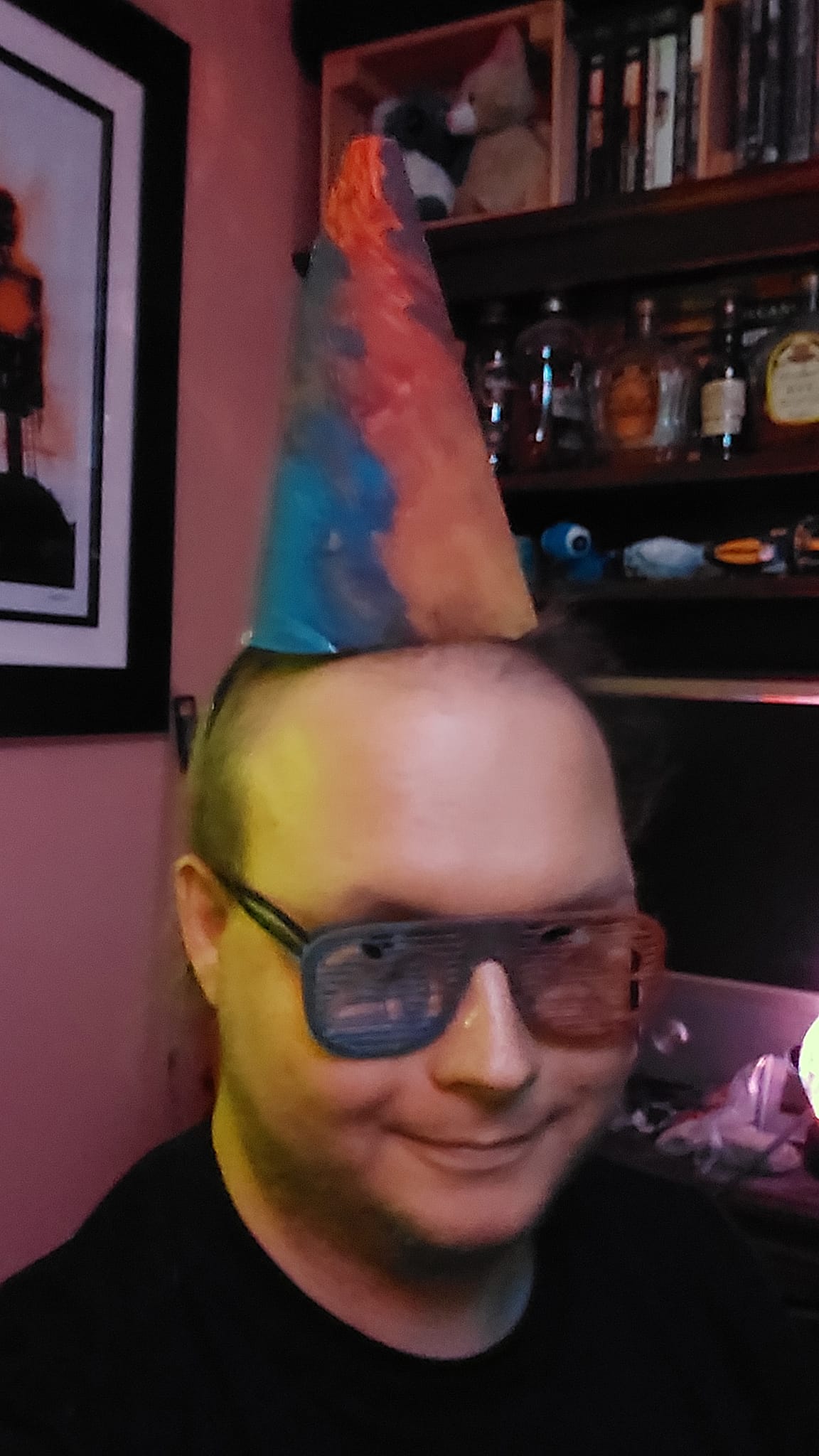 A man sits in a corner, wearing both the hat and painted glasses, the lighting is purple